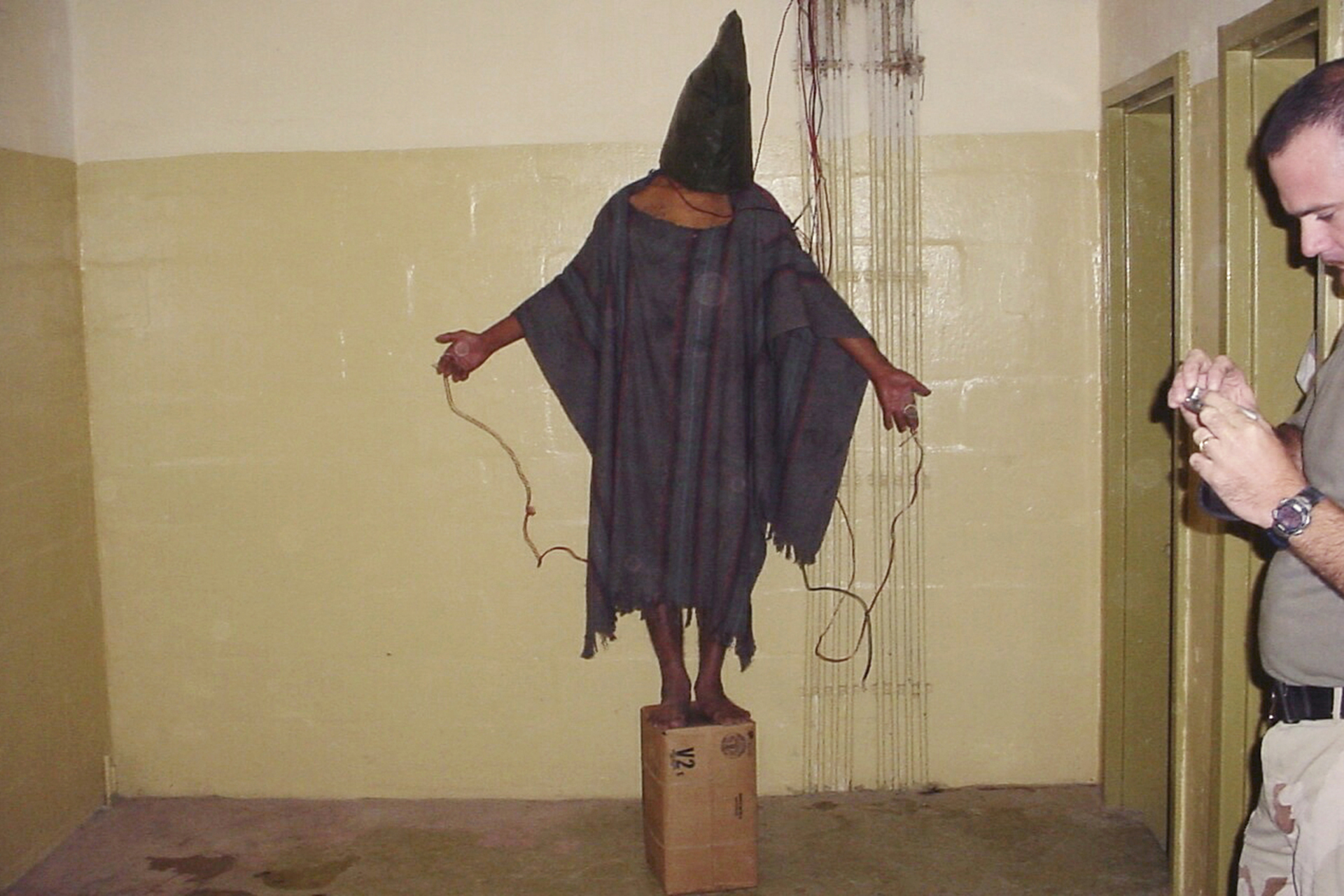 Detenido no identificado se encuntra parado sobre una baja con una bolsa en la cabeza y cables pegados a él para ser torturado en la prisión de Abu Ghraib. (AP)